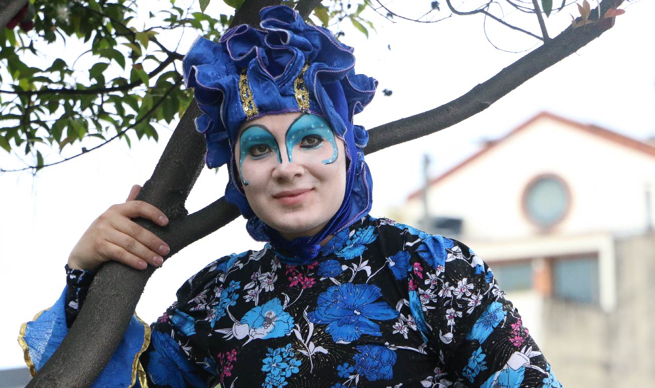 Mujer personificado con un traje azul, cara completamente blanca con iluminación de los ojos hasta las cejas azul para intensificar la mirada y una especie de turbante azul. Paisaje ambiente. 