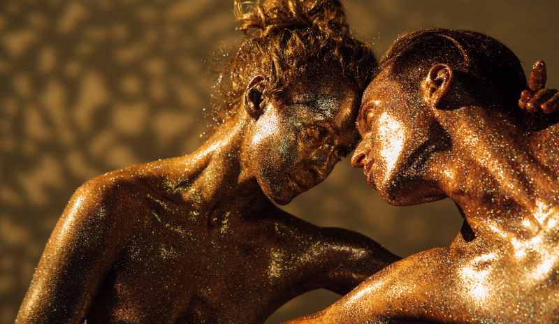 Pareja (hombre y mujer) cubierta con escarcha, sobre su cuerpo desnudo. Tienen el cabello recogido y sus cuerpos se encuentran entrelazados mientras se miran fijamente y sus frentes están juntas. El hombre tie su brazo frente al cuerpo de la mujer.