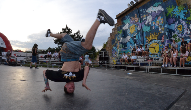 Joven haciendo una pirueta de hip hop. Tiene su cabeza sobre el piso y los pies en el aire, alineados verticalmente con su cabeza. cuenta con camseta negra y jeans. Se ubica sobre una tarima y esta rodeado de un espacio con graffitis.