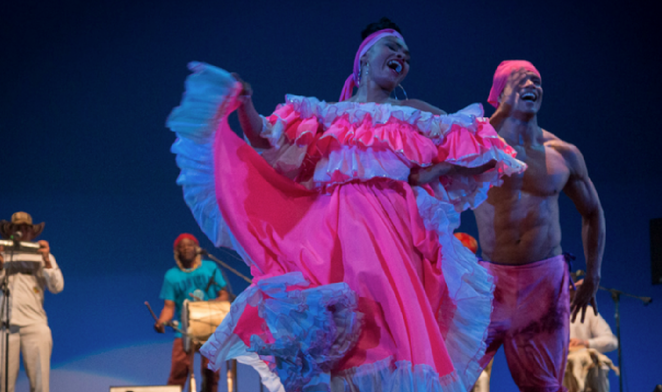Pareja de baile interprentando un bullerengue. La mujer tiene un traje rosado y pañoleta rosada al igual que el hombre, el cual solo tiene un pantalón del mismo color. De fondo músicos en vivo.