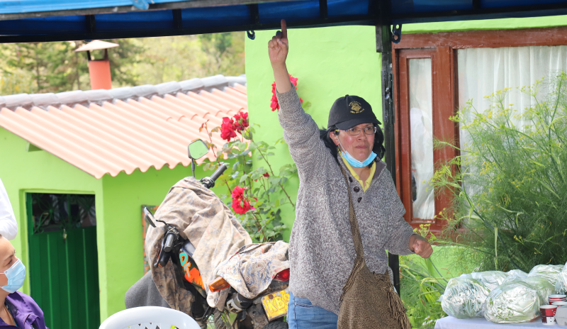Mujer rural con su mano derecha arriba. Se evdencia que hace parte de un círculo de personas que a diferenciia suya se encuentran sentados. 