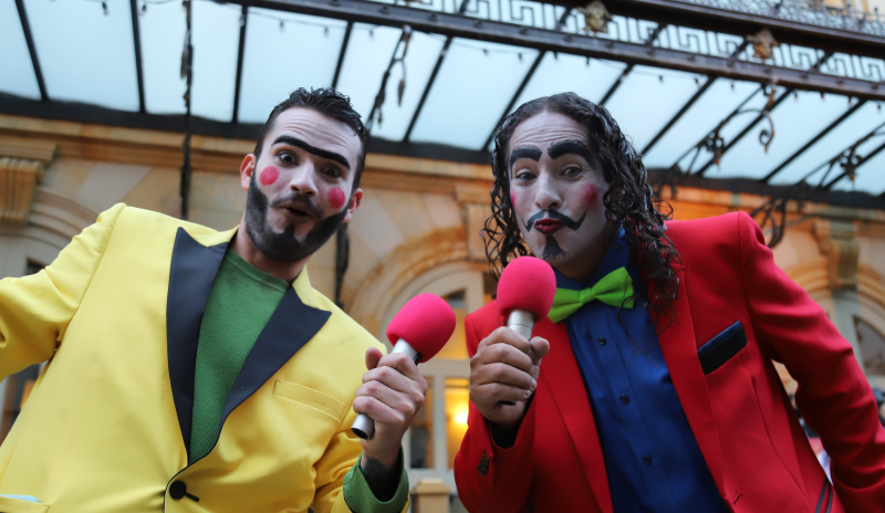 Dos actores: uno con chaqueta roja, camisa azul y corbatín verde. De cabello sobre el hombro, negro rizado (lado derecho) y el otro de chaqueta amarilla con camisa verde (lado izquierdo). Ambos tienen la cara pintada de color blanco. Ambos tienen sobre las mejillas un círculo de color rojo y cuentan con un micrófono de cabeza roja.  