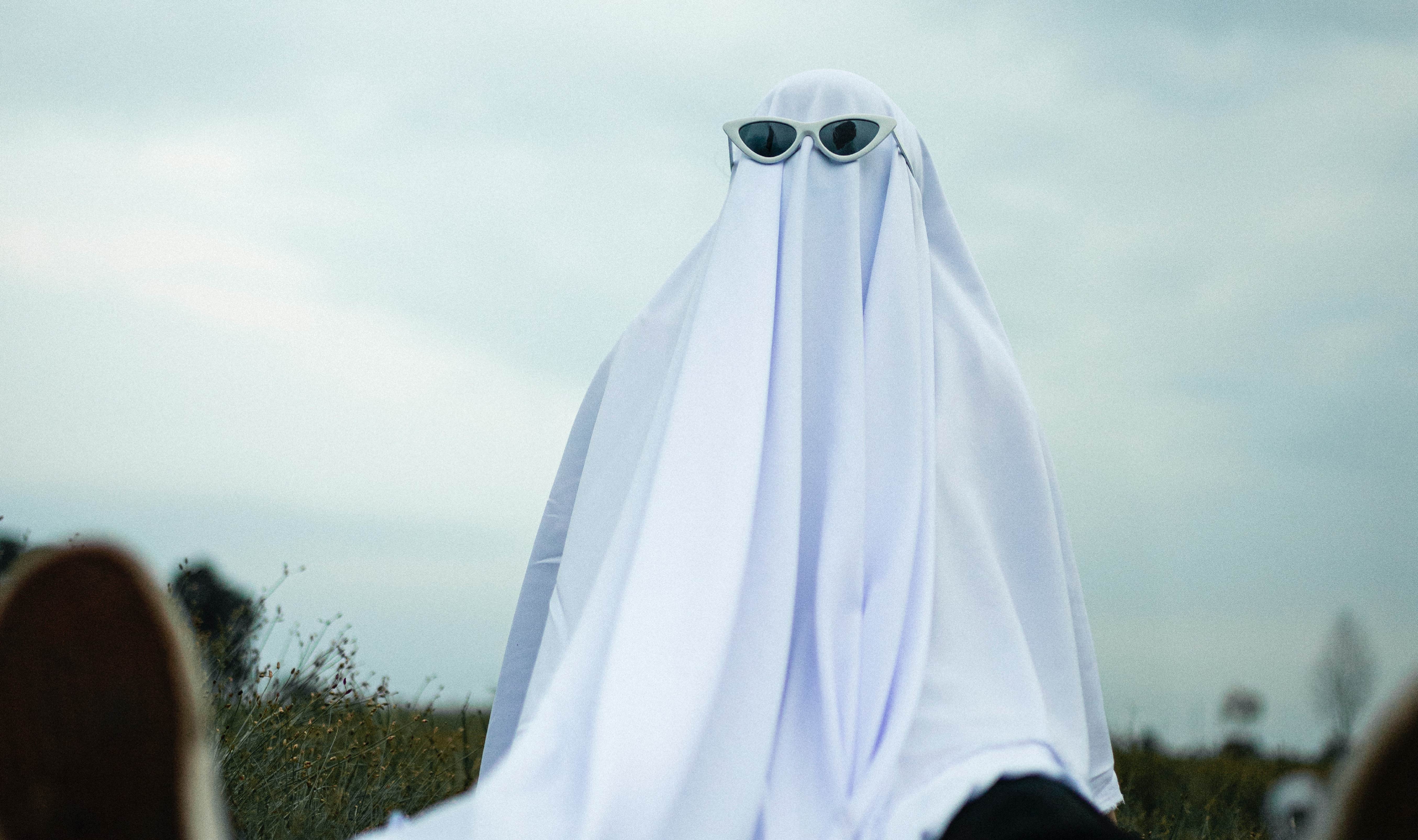 Persona con un traje de fantasma. Tiene unas gafas negras, una tela blanca que lo cubre por completo, pantalones oscuros, y tenis. Se encuentra sentado al aire libre. 