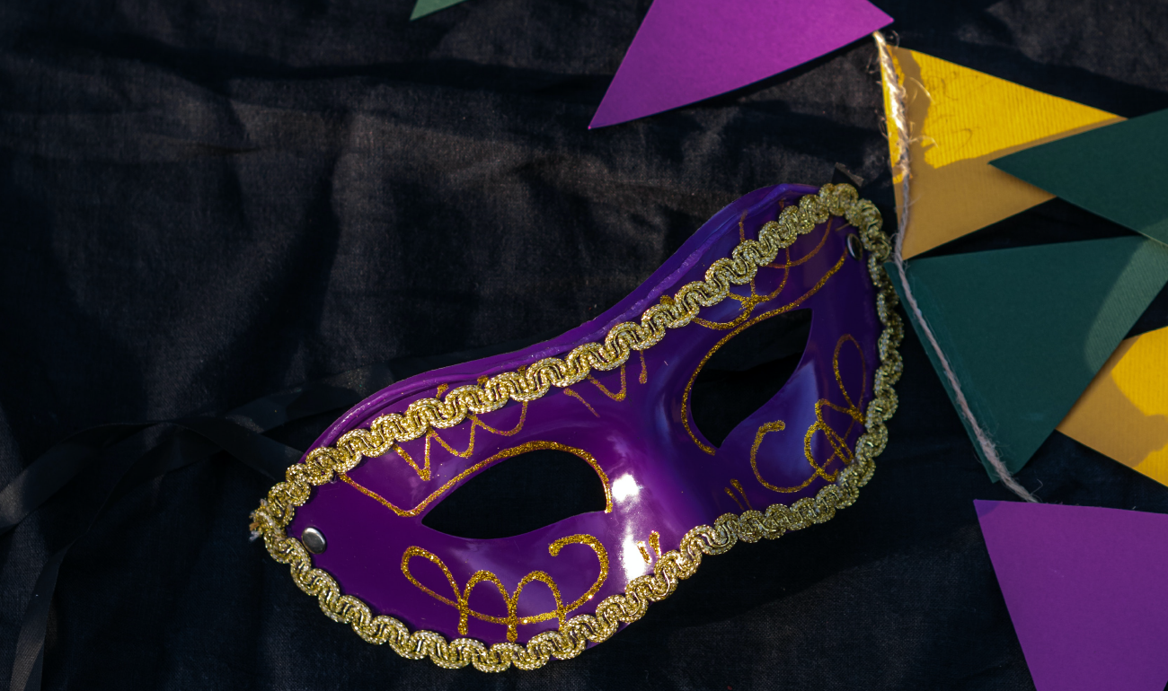Antifaz morado con bordes dorados sobre una tela negra. Al lado derecho se ven banderines morados, amarillos y verdes