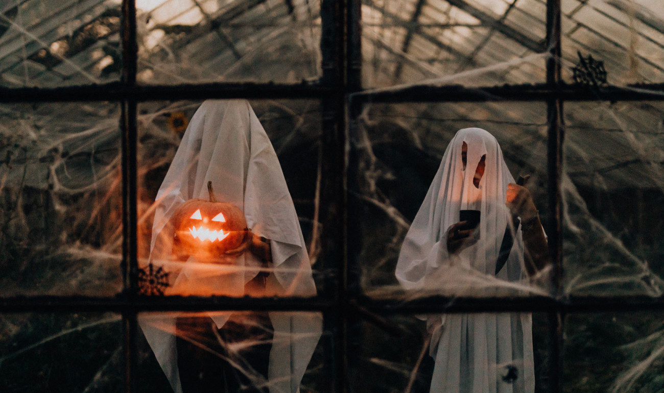 Dos personas con traje de fantasma tras una estructura de ventanas. La persona de la izquierda tiene una calabaza con luz en las manos.