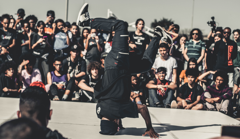 Joven haciendo una pirueta de hip hop. Tiene su cabeza sobre el piso y los pies en el aire, alineados verticalmente con su cabeza. cuenta con camseta negra y jeans. Se ubica sobre una tarima y esta rodeado de un espacio con graffitis.